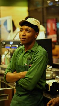 Barista in a coffee shop wearing a green apron and cap, leaning on the counter.